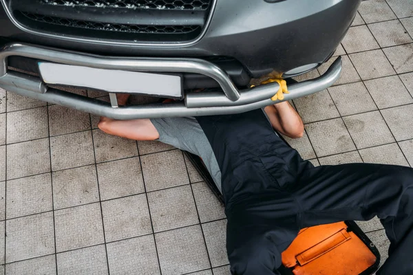 Vue grand angle de l'ouvrier couché et travaillant sous la voiture dans l'atelier de mécanique — Photo de stock