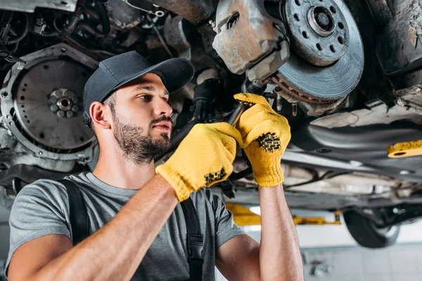 Professional mechanic in uniform repairing car without wheel in workshop — Stock Photo