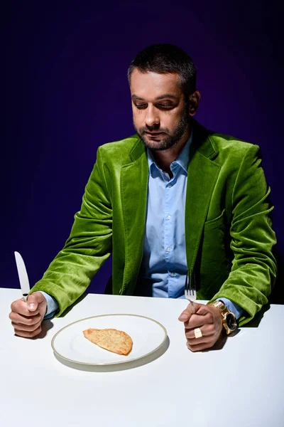 Homme en veste de velours vert élégant assis à table avec de la pâtisserie de viande sur plaque avec fond bleu derrière — Photo de stock