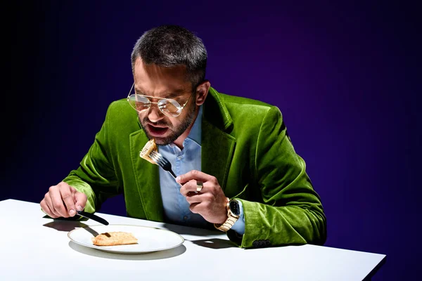 Uomo in elegante giacca di velluto verde mangiare cheburek carne a tavola con sfondo blu dietro — Foto stock