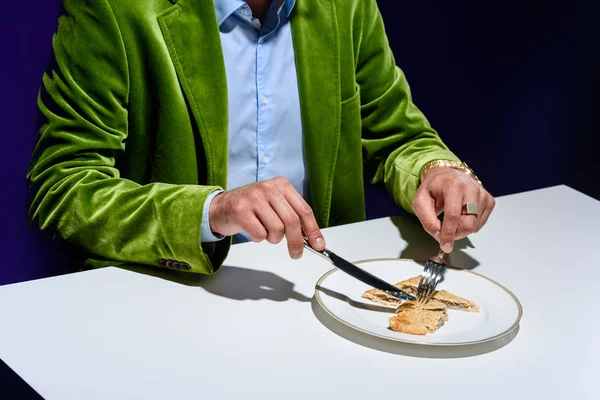 Vue partielle de l'homme en veste de velours vert élégant découpe pâtisserie de viande sur plaque avec fond bleu derrière — Photo de stock