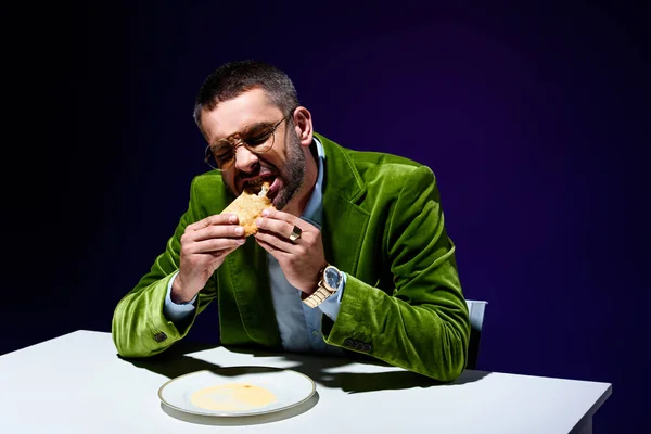Retrato de homem na elegante jaqueta de veludo verde comendo massa de carne à mesa com fundo azul atrás — Fotografia de Stock