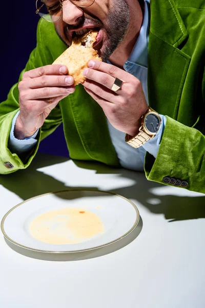 Cropped shot of man in stylish green velvet jacket eating cheburek at table with blue background behind — Stock Photo