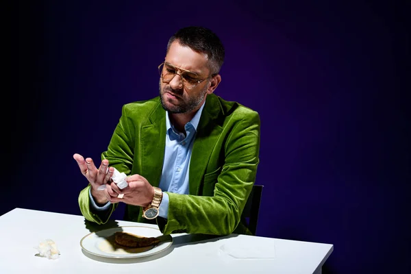 Man in stylish green velvet jacket drying hands with napkin at table with meat pastry on plate with blue background behind — Stock Photo