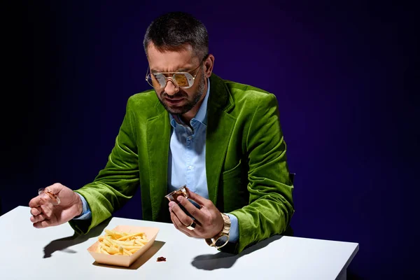 Retrato del hombre en ropa de moda sentado a la mesa con papas fritas y ketchup con fondo azul - foto de stock