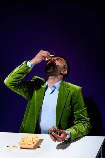 Retrato de homem elegante em casaco de veludo verde comendo batatas fritas com ketchup à mesa com fundo azul — Fotografia de Stock