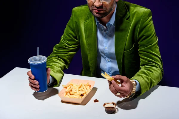 Vista parziale dell'uomo in abiti alla moda con bibita gassata seduto a tavola con patatine fritte e ketchup con sfondo blu — Foto stock