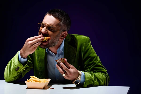 Homem elegante em casaco de veludo comendo batatas fritas com ketchup à mesa com fundo azul — Fotografia de Stock
