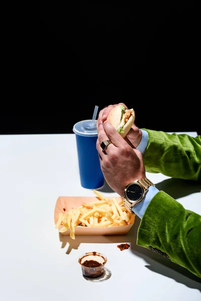 Частковий вигляд людини в оксамитовій куртці з бургер, що сидить за столом з картоплею фрі та содовою на чорному тлі — стокове фото