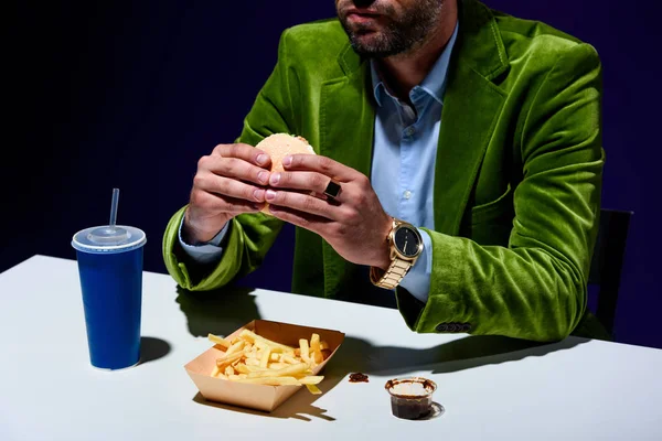 Tiro recortado de hombre en chaqueta de terciopelo comiendo hamburguesa en la mesa con gritos franceses y bebida de soda con fondo azul - foto de stock