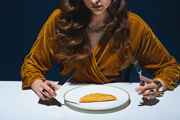 Cropped shot of woman in luxury clothing sitting at table with meat pastry on plate with blue backdrop — Stock Photo