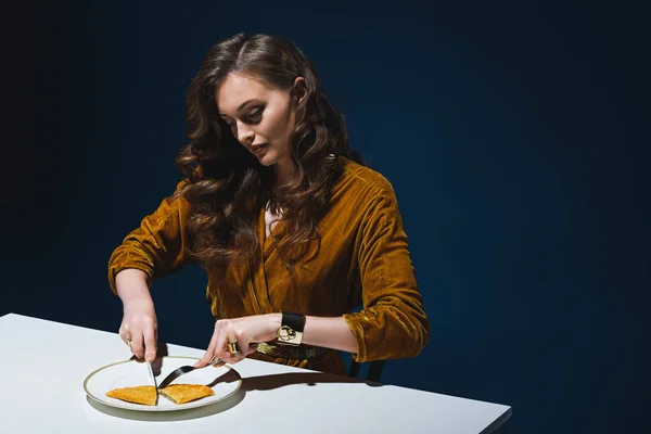 Woman in stylish clothing cutting unhealthy cheburek at table with blue backdrop behind — Stock Photo