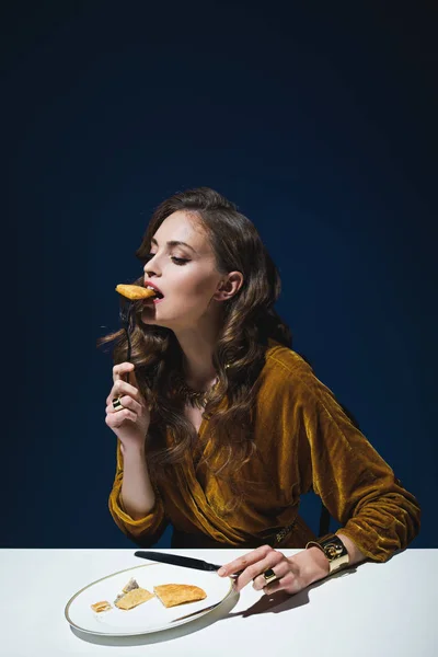 Belle femme en vêtements élégants manger de la pâtisserie de viande à la table avec fond bleu — Photo de stock