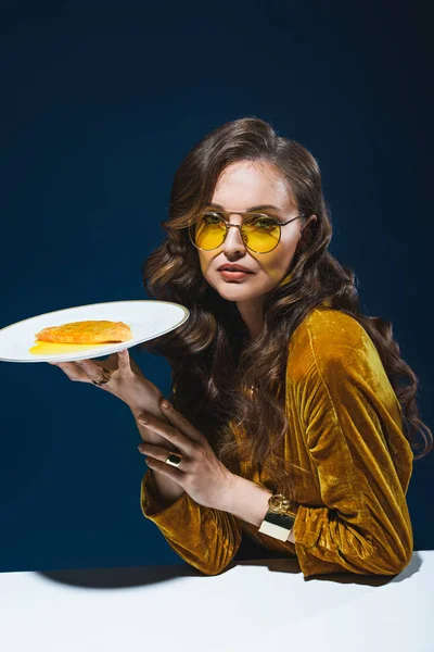 Portrait of beautiful woman in stylish clothing with unhealthy cheburek on plate sitting at table with blue background behind — Stock Photo