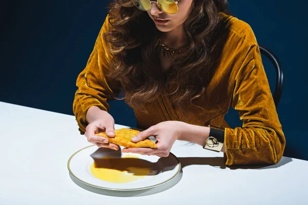 Partial view of fashionable woman with unhealthy meat pastry sitting at table with blue backdrop — Stock Photo