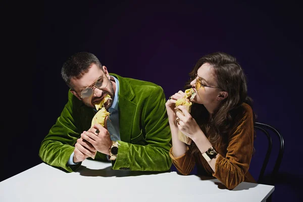 Couple in luxury velvet clothing eating shawarma while sitting at table with dark background behind — Stock Photo