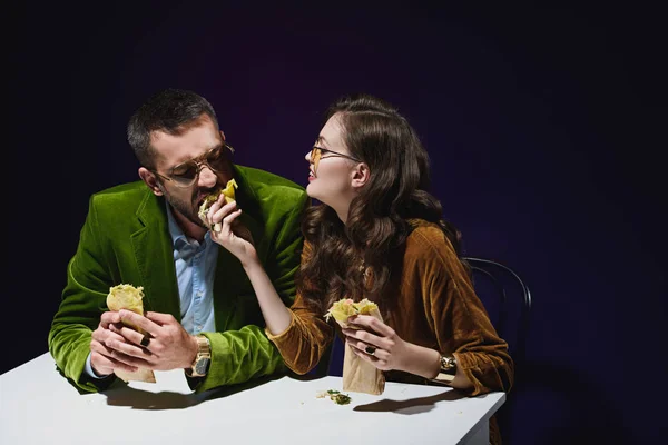 Casal em roupas de veludo de luxo com shawarma sentado à mesa com fundo escuro atrás — Fotografia de Stock