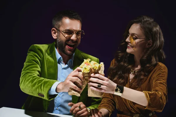 Retrato de pareja sonriente en ropa de terciopelo de lujo con shawarma sentado en la mesa con fondo oscuro detrás - foto de stock