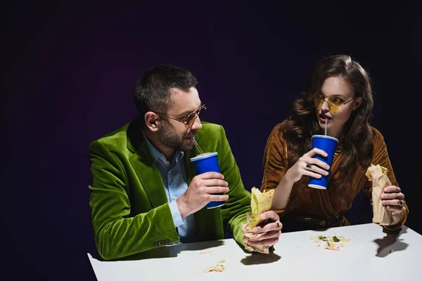 Retrato de pareja en ropa de terciopelo de lujo con shawarma y bebidas gaseosas sentado a la mesa con fondo oscuro detrás - foto de stock