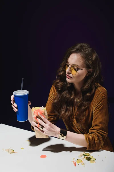 Portrait de femme séduisante en vêtements à la mode avec soda et shawarma assis à table avec fond bleu — Photo de stock