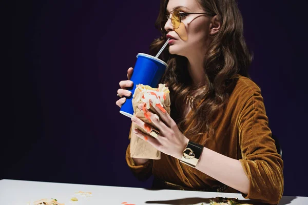 Vista lateral da mulher atraente em roupas da moda com bebida de refrigerante e shawarma sentado à mesa com fundo azul — Fotografia de Stock