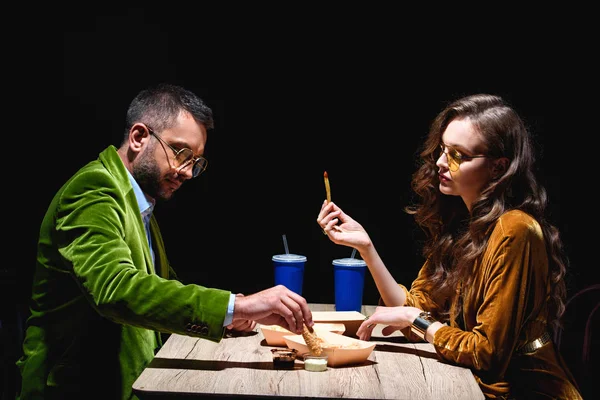 Vista lateral de la pareja en ropa de terciopelo elegante sentado a la mesa con anillos de cebolla frita, papas fritas y salsas con fondo negro — Stock Photo