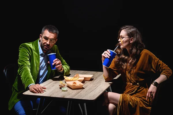 Vue latérale du couple dans des vêtements élégants en velours assis à table avec des rondelles d'oignons frits, des frites et des sauces avec fond noir — Photo de stock