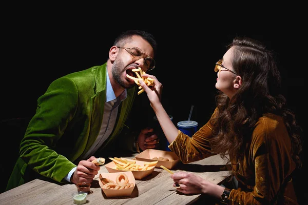 Vista laterale di coppia in abiti di velluto elegante mangiare patatine fritte, anelli di cipolla fritti e salse a tavola con sfondo nero — Foto stock