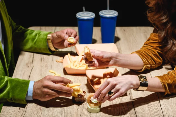 Corte tiro de casal em roupas de veludo elegante sentado à mesa com anéis de cebola frita, batatas fritas e molhos com fundo preto — Fotografia de Stock