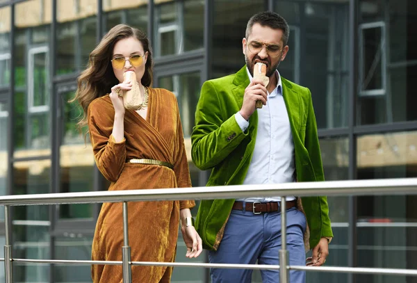 Porträt eines Paares im Luxus-Outfit, das französische Hotdogs isst, während es auf der Straße spaziert — Stockfoto