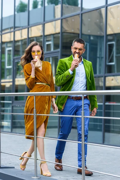 Couple en tenue de luxe manger des hot-dogs français tout en marchant dans la rue — Photo de stock