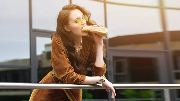 Porträt einer attraktiven Frau in luxuriöser Kleidung und Sonnenbrille, die auf der Straße französischen Hot Dog isst — Stockfoto