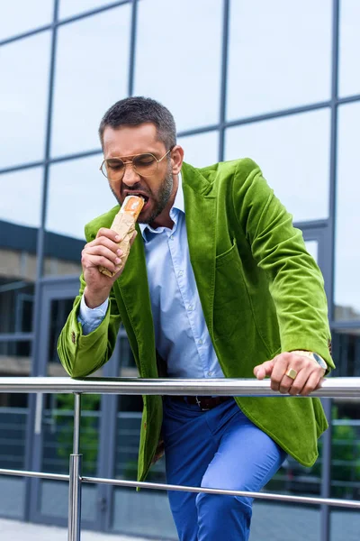 Portrait of man in green velvet jacket eating french hot dog on street — Stock Photo