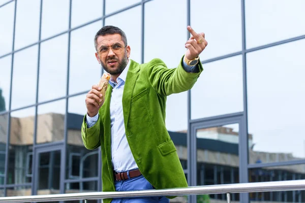 Portrait of man in green velvet jacket with french hot dog in hand showing middle finger on street — Stock Photo
