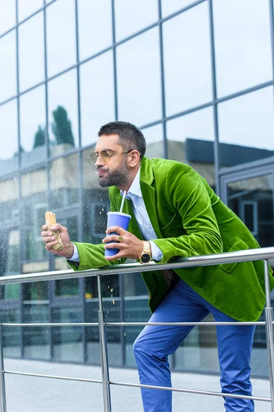 Side view of man in green velvet jacket with french hot dog and soda drink in hands on street — Stock Photo