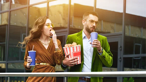 Retrato de pareja de moda en ropa de terciopelo con bebidas gaseosas comiendo patas de pollo fritas mientras camina por la calle - foto de stock