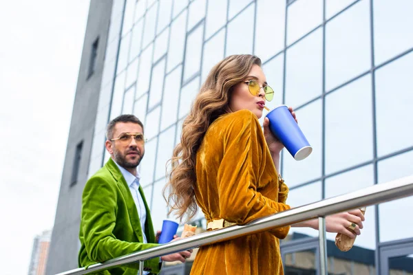 Vista lateral de pareja elegante en ropa de terciopelo con bebidas gaseosas y perros calientes franceses en la calle - foto de stock