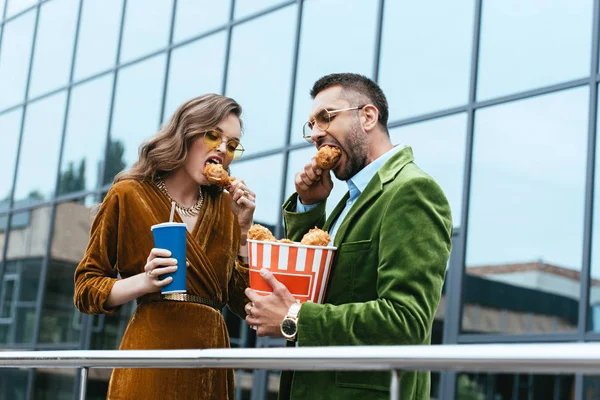 Portrait of fashionable couple in velvet clothing eating fried chicken legs on street — Stock Photo