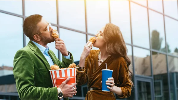 Side view of fashionable couple in velvet clothing eating fried chicken legs on street — Stock Photo