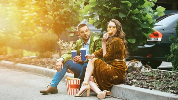 Couple à la mode en vêtements de velours avec des boissons manger des jambes de poulet frit dans la rue — Photo de stock