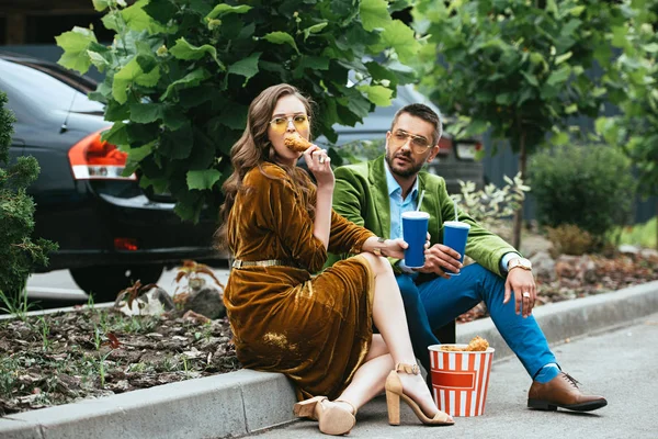 Coppia alla moda in abiti di velluto con gambe di pollo fritto e bevande in strada — Foto stock