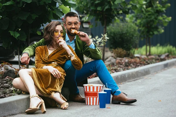 Модная пара в бархатной одежде ест жареных куриных ножек на улице — стоковое фото