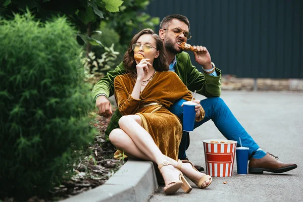 Pareja de moda en ropa de terciopelo comiendo patas de pollo frito en la calle - foto de stock