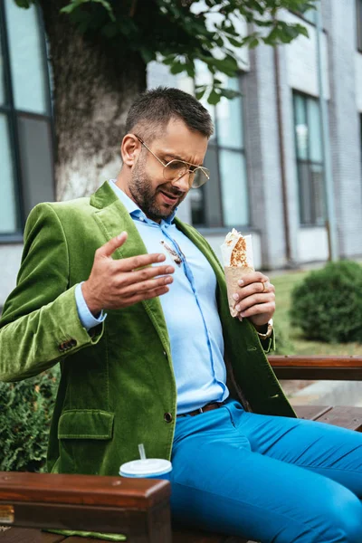 Homme à la mode en chemise sale avec shawarma à la main assis sur le banc sur la rue — Photo de stock