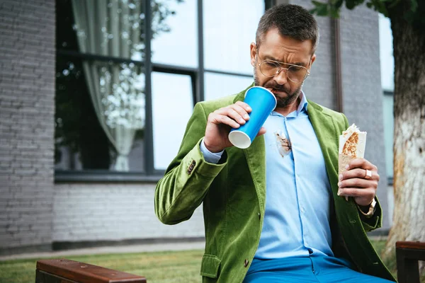 Portrait of man in fashionable clothing with shawarma in hand drinking soda drink while sitting on bench on street — Stock Photo