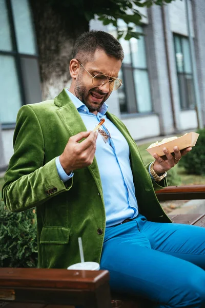 Portrait of emotional man in green velvet jacket with ketchup on shirt and french fries in hands sitting on bench on street — Stock Photo