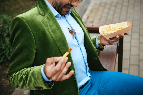 Partial view of man in green velvet jacket with ketchup on shirt and french fries in hands sitting on bench on street — Stock Photo