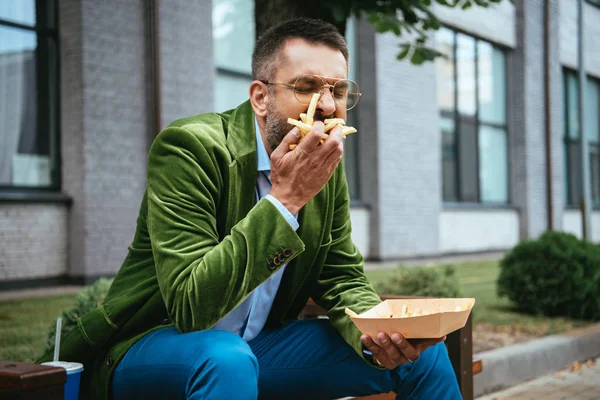 Portrait d'homme en veste de velours vert mangeant des frites assis sur un banc dans la rue — Photo de stock