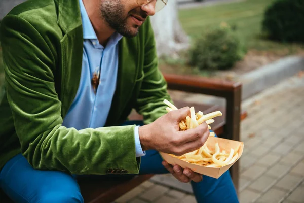 Vue partielle de l'homme en velours vert veste avec des frites assis sur le banc sur la rue — Photo de stock