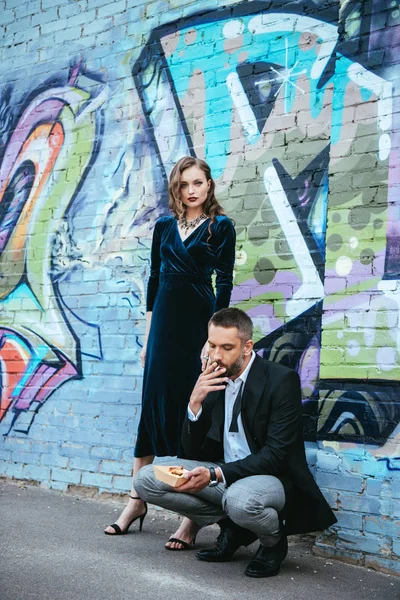 Pareja en traje de moda con papas fritas cerca de la pared con graffiti en la calle - foto de stock